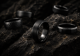 a collection of mens black wedding bands photographed on a dark stone surface