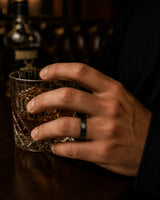 Man wearing a black beveled whiskey barrel wood wedding ring while holding a crystal glass of whiskey in a dark cinematic bar setting.