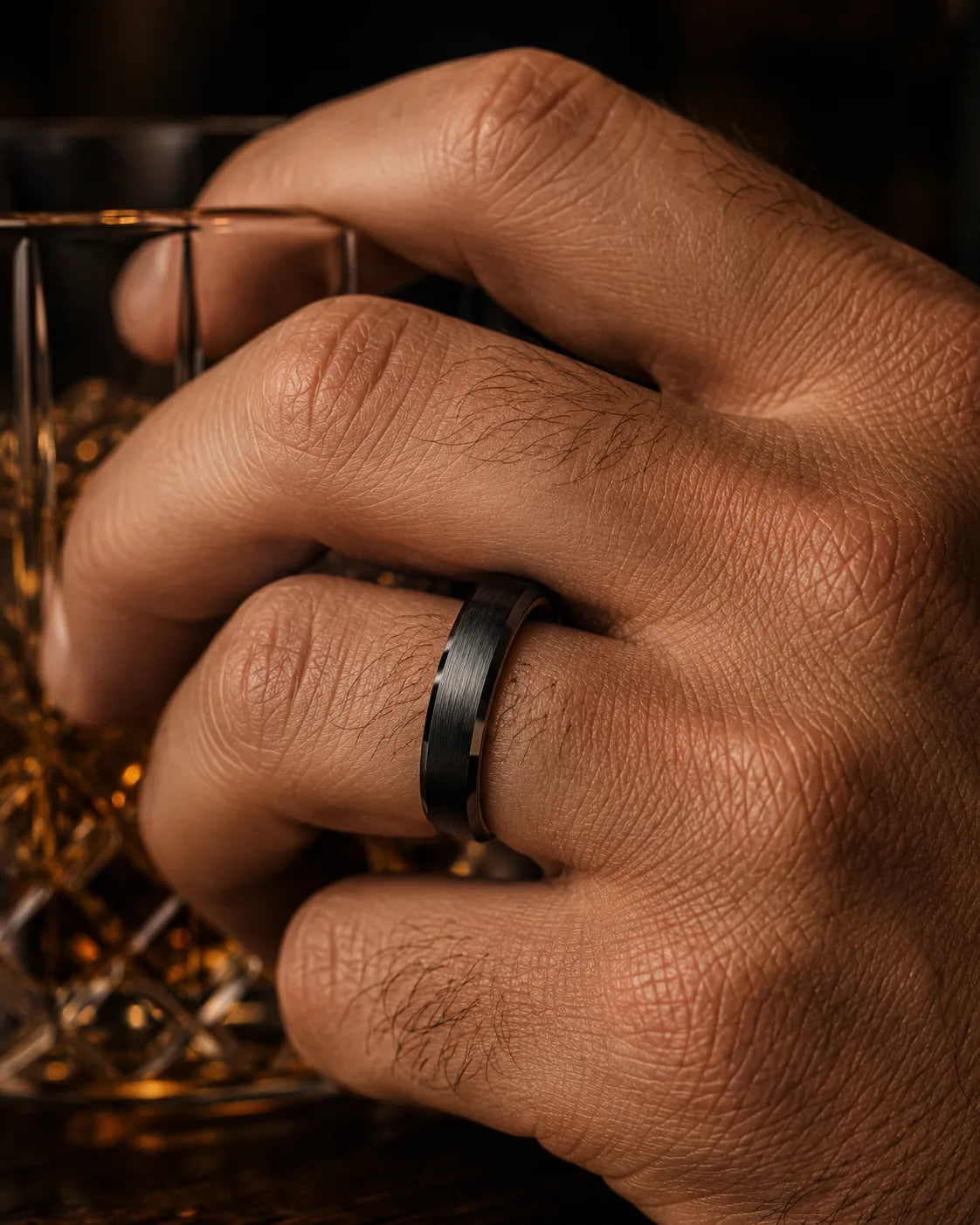 Macro close up of a black beveled whiskey barrel wood wedding ring worn on a man's finger while holding a crystal whiskey tumbler.