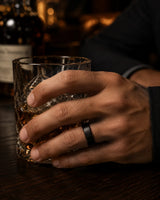 Man wearing an 6mm brushed black tungsten wedding ring with polished edges and genuine whiskey wood sleeve while holding a crystal whiskey glass.