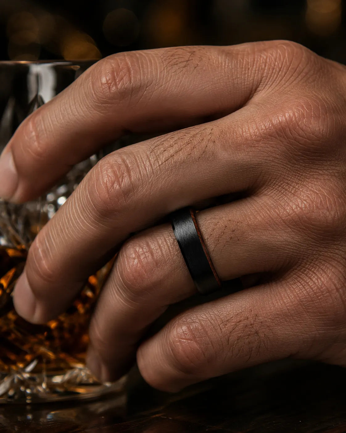 Man wearing an 6mm brushed black tungsten wedding ring with polished edges and genuine whiskey wood sleeve while holding a crystal whiskey glass.