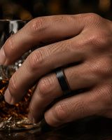Man wearing an 6mm brushed black tungsten wedding ring with polished edges and genuine whiskey wood sleeve while holding a crystal whiskey glass.