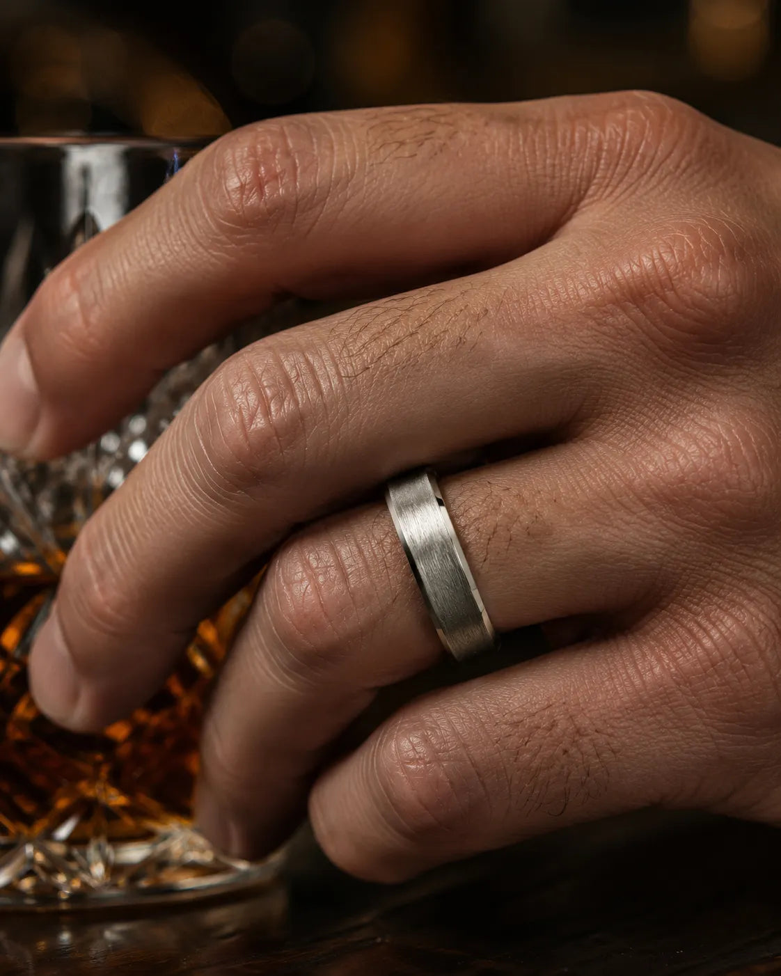 Close up of an 6mm brushed silver tungsten wedding ring with polished edges and genuine maple wood sleeve worn while holding a crystal whiskey tumbler.