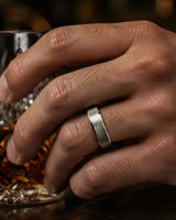 Close up of an 6mm brushed silver tungsten wedding ring with polished edges and genuine maple wood sleeve worn while holding a crystal whiskey tumbler.