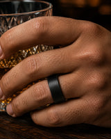 Close up of a man wearing a brushed black tungsten wedding ring with a maple wood sleeve interior while holding a whiskey glass.