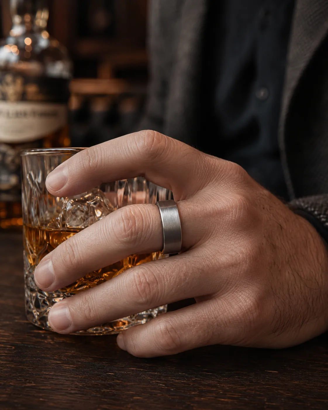 Man wearing a brushed silver tungsten wedding ring with a maple wood sleeve interior while holding a crystal whiskey tumbler in a luxury whiskey bar.