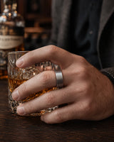 Man wearing a brushed silver tungsten wedding ring with a maple wood sleeve interior while holding a crystal whiskey tumbler in a luxury whiskey bar.