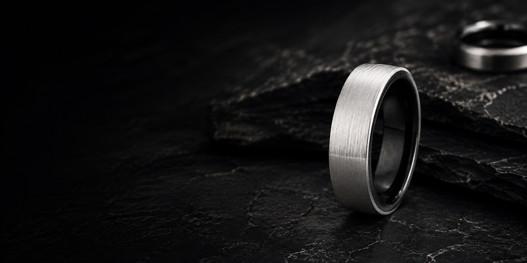 Luxury brushed silver and black tungsten wedding ring photographed on dark slate with moody industrial lighting