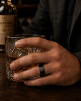 Man wearing a brushed black tungsten wedding ring with a maple wood sleeve interior while holding a crystal whiskey tumbler in a luxury whiskey bar.