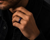 Close-up lifestyle photograph of a man wearing a black tungsten wedding ring with purple amethyst stone inlay in a moody modern editorial setting.