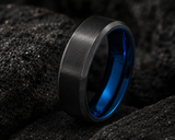 Macro close-up of a black tungsten men’s wedding ring with blue interior resting on rugged charcoal volcanic stone.