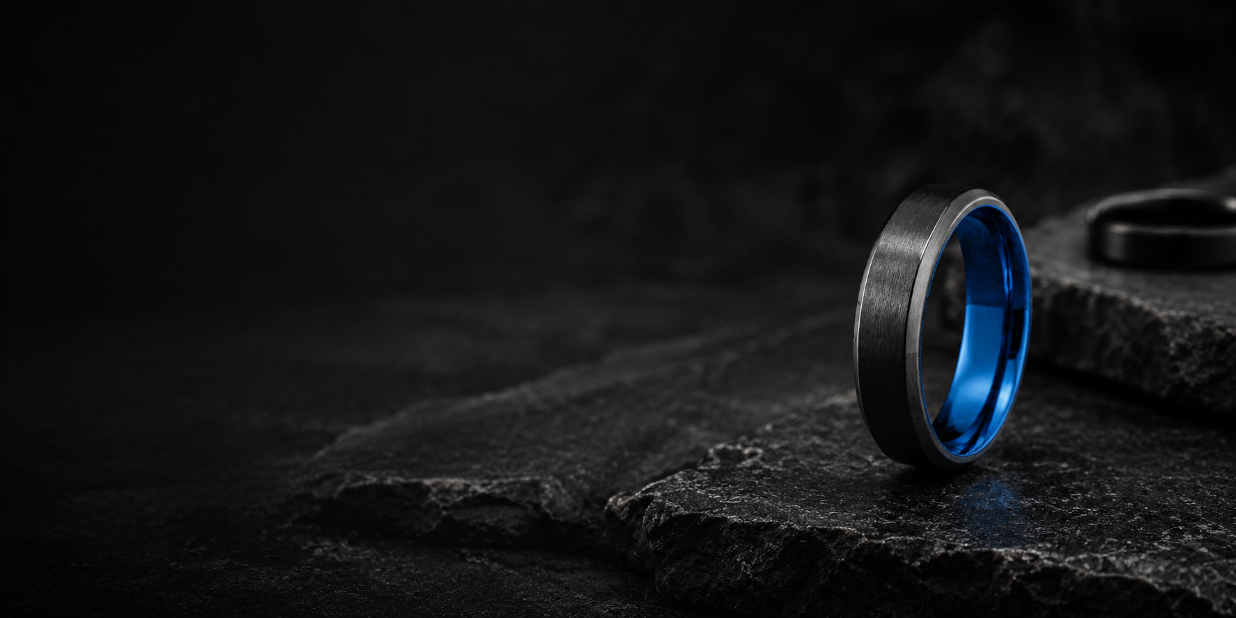 Premium editorial photograph of a black brushed tungsten wedding band with blue inner sleeve in a moody masculine setting.