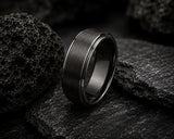 Macro close-up of a black brushed men’s wedding ring resting on volcanic rock with detailed texture and moody industrial lighting.
