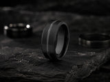 Black groove tungsten wedding ring on dark volcanic rock with moody cinematic lighting and rugged charcoal slate textures.