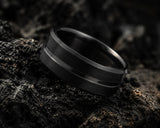 Ultra close-up macro photograph of a black groove tungsten wedding ring showing brushed texture and polished details on volcanic rock background.