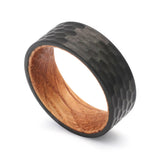 Angled view of a black hammered tungsten whiskey barrel wedding ring with a real oak wood interior.