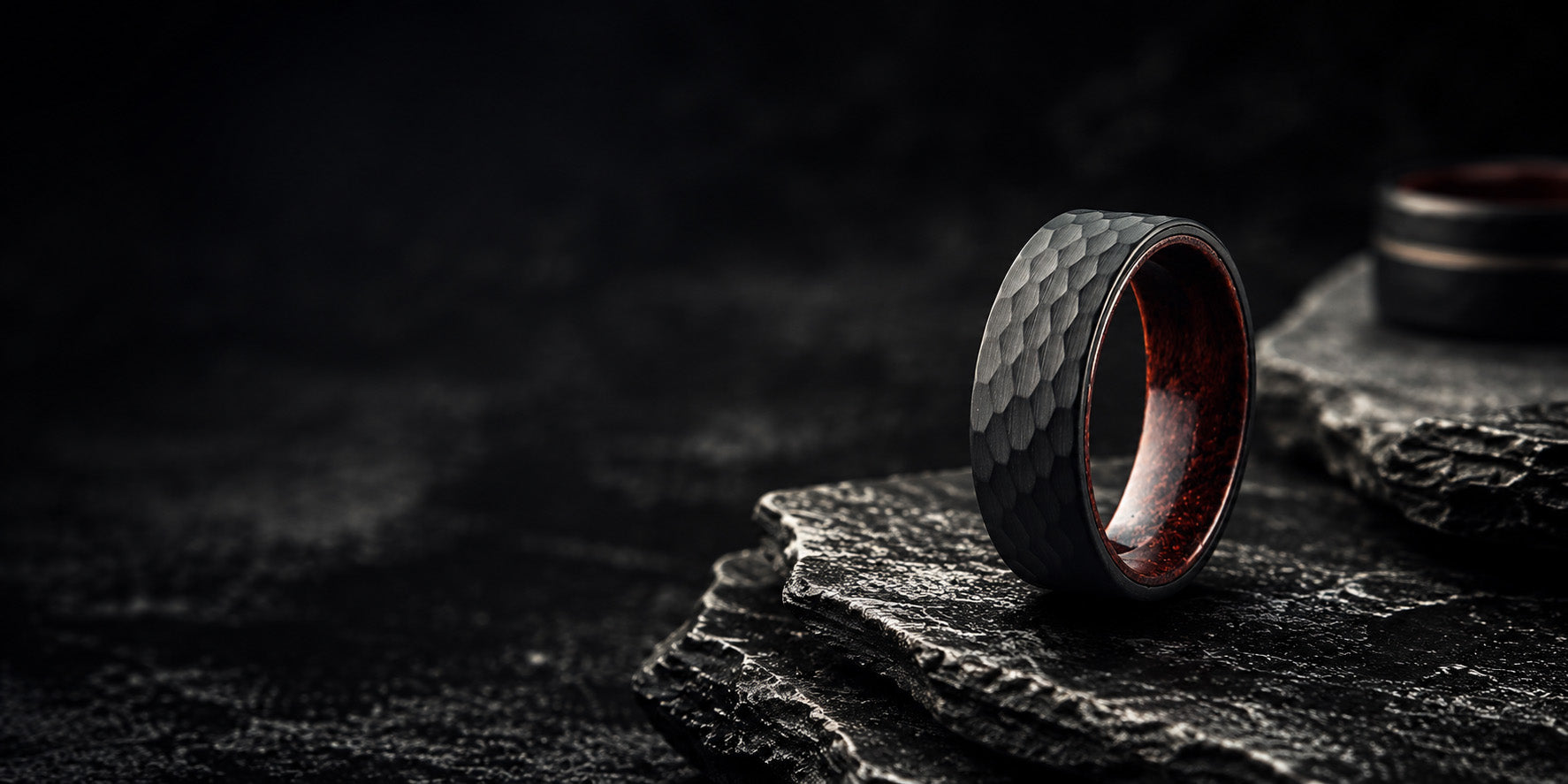 Black hammered men’s wedding ring with rich wood interior resting on dark volcanic stone in a premium cinematic ecommerce hero image.