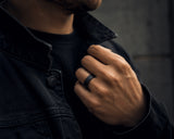 Male hand wearing a black hammered wedding ring with wood interior styled with dark modern clothing in an urban luxury lifestyle setting.