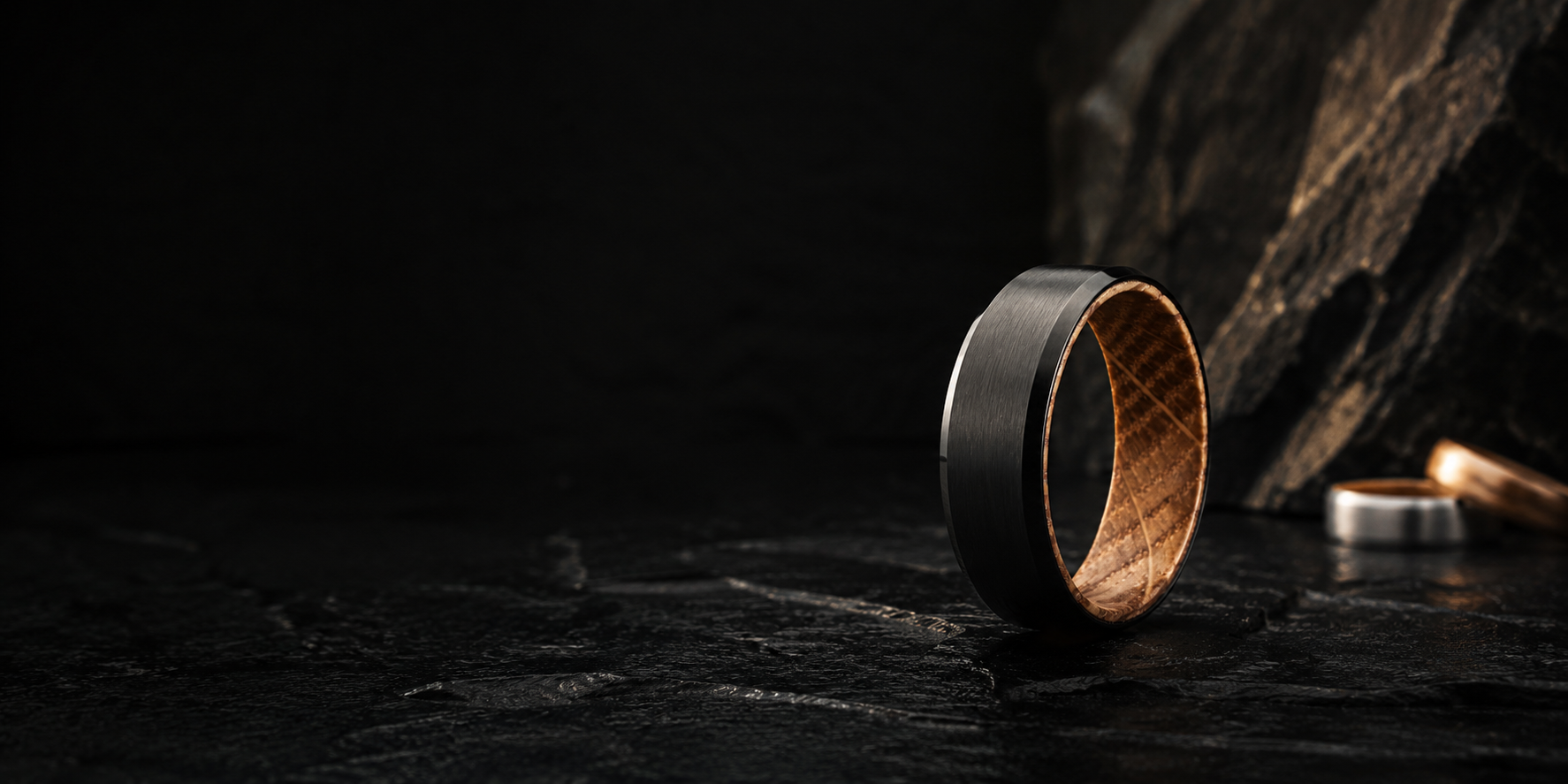 Black tungsten men’s wedding ring with natural oak wood interior on dark volcanic slate background in cinematic luxury product photography style.