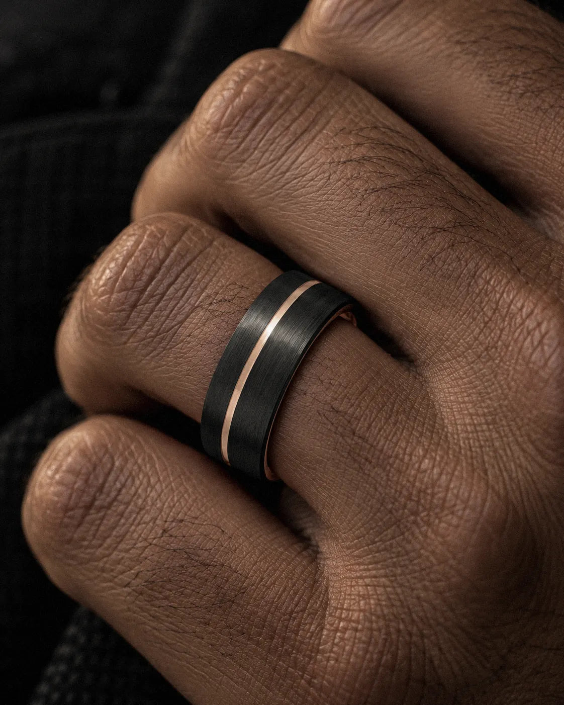 Close-up of a brushed black men’s wedding ring with a rose gold offset groove worn on the ring finger.