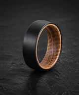 Black whiskey barrel wedding ring with a reclaimed oak wood interior on a dark stone background.