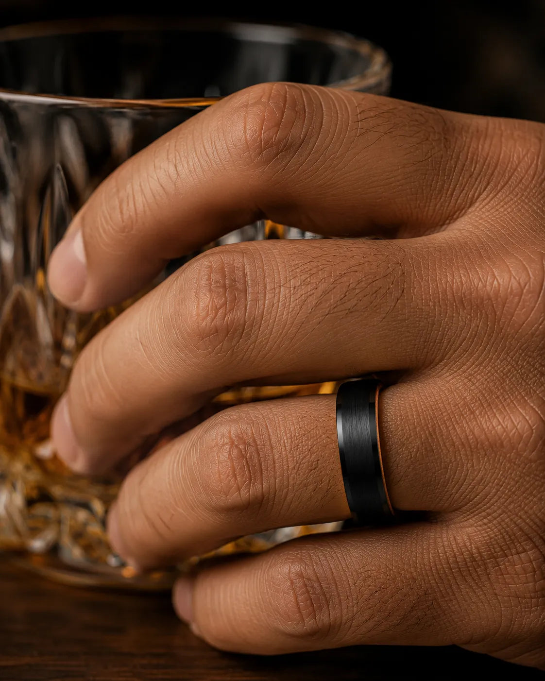 Macro close up of a black brushed tungsten wedding ring with polished beveled edges and a whiskey barrel wood interior worn while holding a crystal whiskey glass.