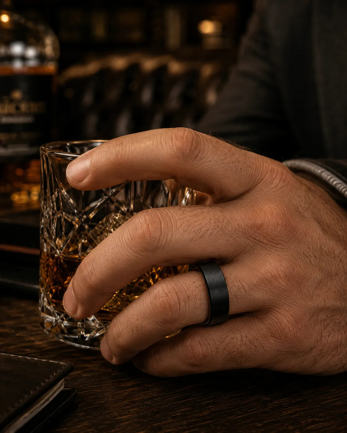 Man wearing a black tungsten wedding ring with a brushed finish and whiskey wood interior while holding a whiskey glass in a dark luxury bar.