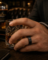 Man wearing a black tungsten wedding ring with a brushed finish and whiskey wood interior while holding a whiskey glass in a dark luxury bar.