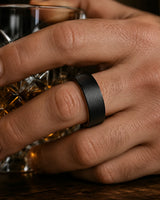 Close-up macro photograph of a black tungsten wedding ring with a brushed finish and whiskey wood interior worn while holding a crystal whiskey tumbler in warm cinematic lighting.