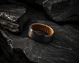 Macro close-up of black tungsten and oak wood wedding ring resting on dark slate stone showing brushed texture and polished edges.