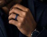 Close-up lifestyle photograph of a man wearing a silver tungsten wedding ring with blue sapphire stone inlay in a moody modern editorial setting.