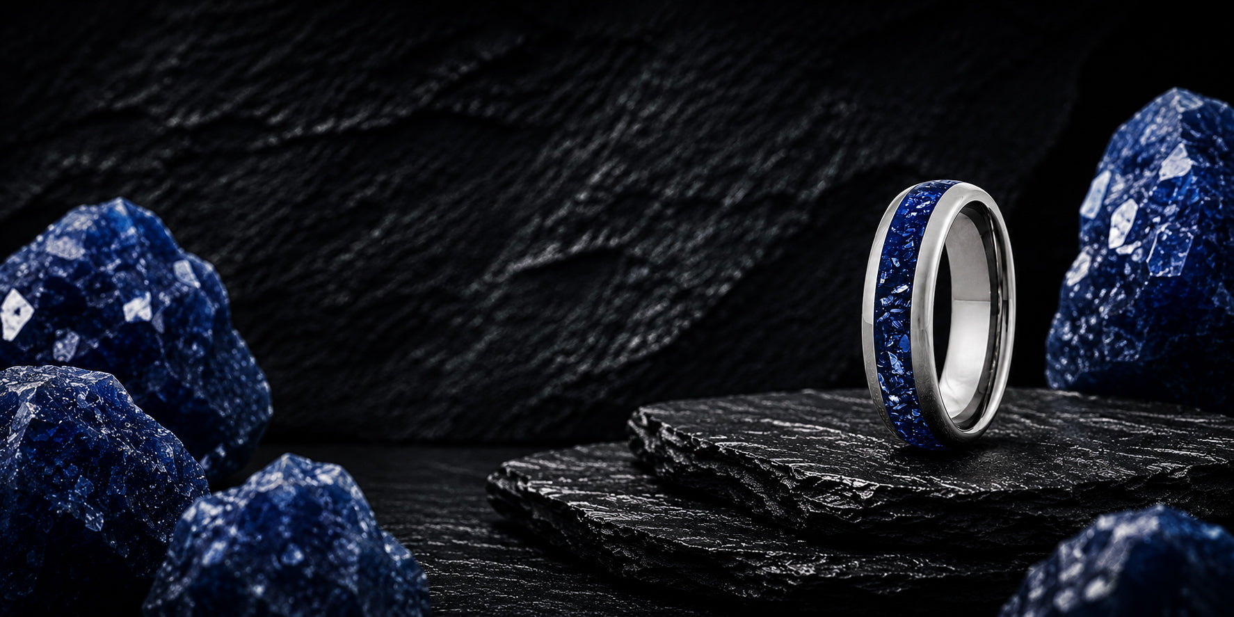 Wide desktop banner for blue sapphire wedding rings collection featuring a silver tungsten ring with vivid blue gemstone inlay positioned on a dark luxury slate background.