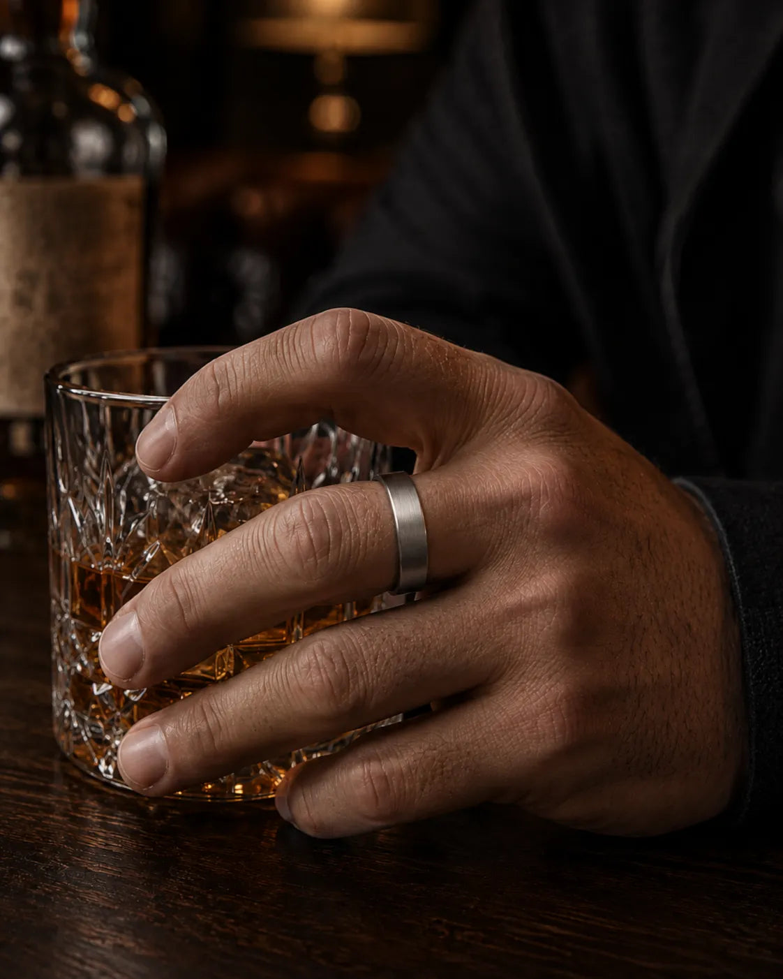 Man wearing a brushed silver tungsten wedding ring with a maple wood sleeve interior while holding a crystal whiskey tumbler in a luxury whiskey bar.