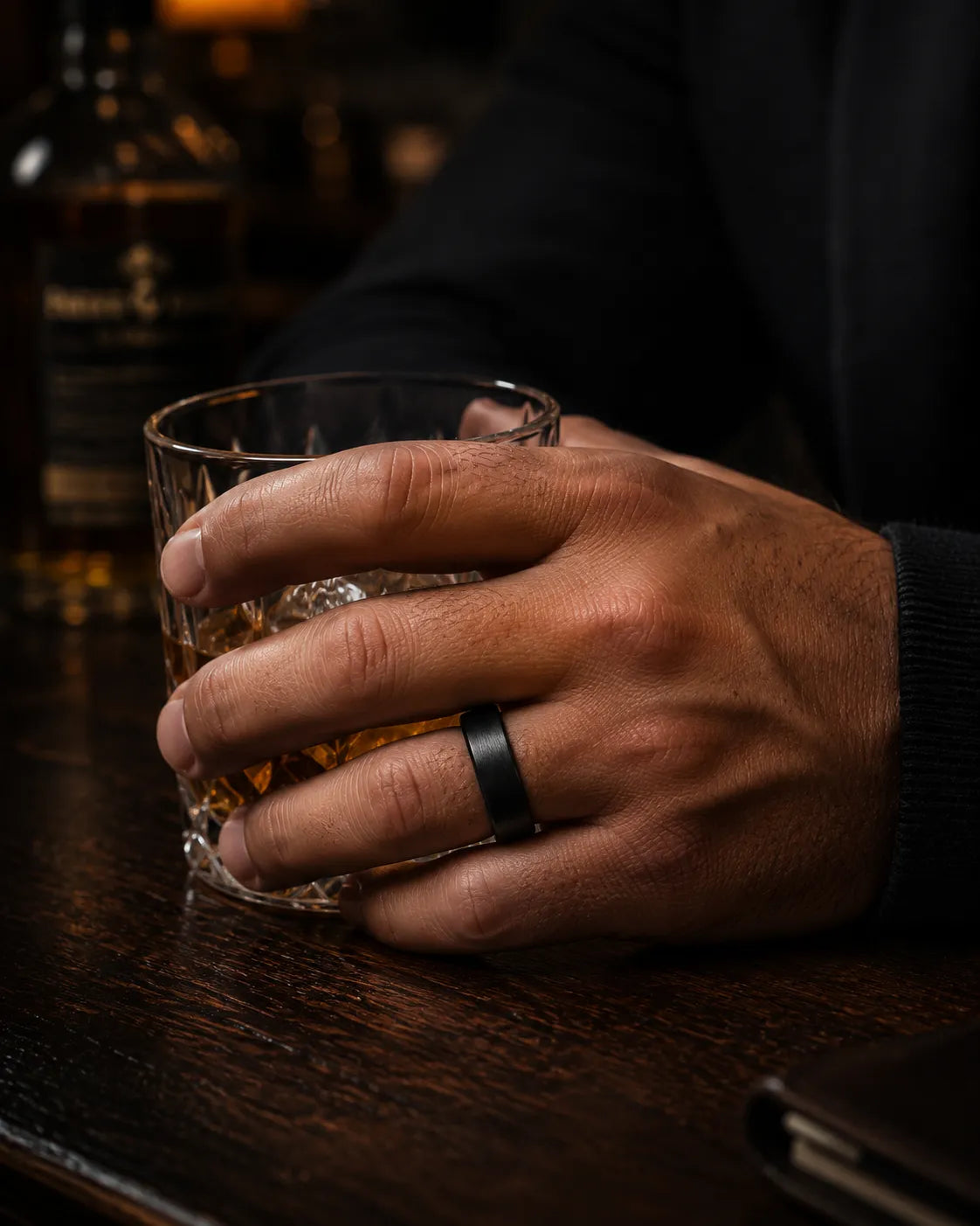 Man wearing a brushed black tungsten wedding ring with a maple wood sleeve interior while holding a crystal whiskey tumbler in a luxury whiskey bar.