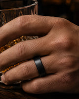 Close up of a man wearing a brushed black tungsten wedding ring with a maple wood sleeve interior while holding a whiskey glass.