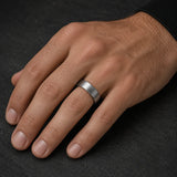 Brushed silver tungsten wedding ring worn on a man's left hand against a dark stone background.