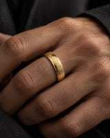 Close up of a polished gold tungsten wedding ring with a smooth domed profile, worn on a man's ring finger against a dark background.