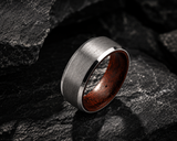 Close-up of brushed silver wedding ring with whiskey barrel wood interior resting on dark stone surface showing textured finish and durable materials
