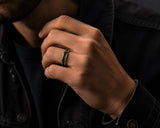 Close-up lifestyle photograph of a man wearing a gold tungsten wedding ring with emerald green inlay in a moody modern editorial setting.