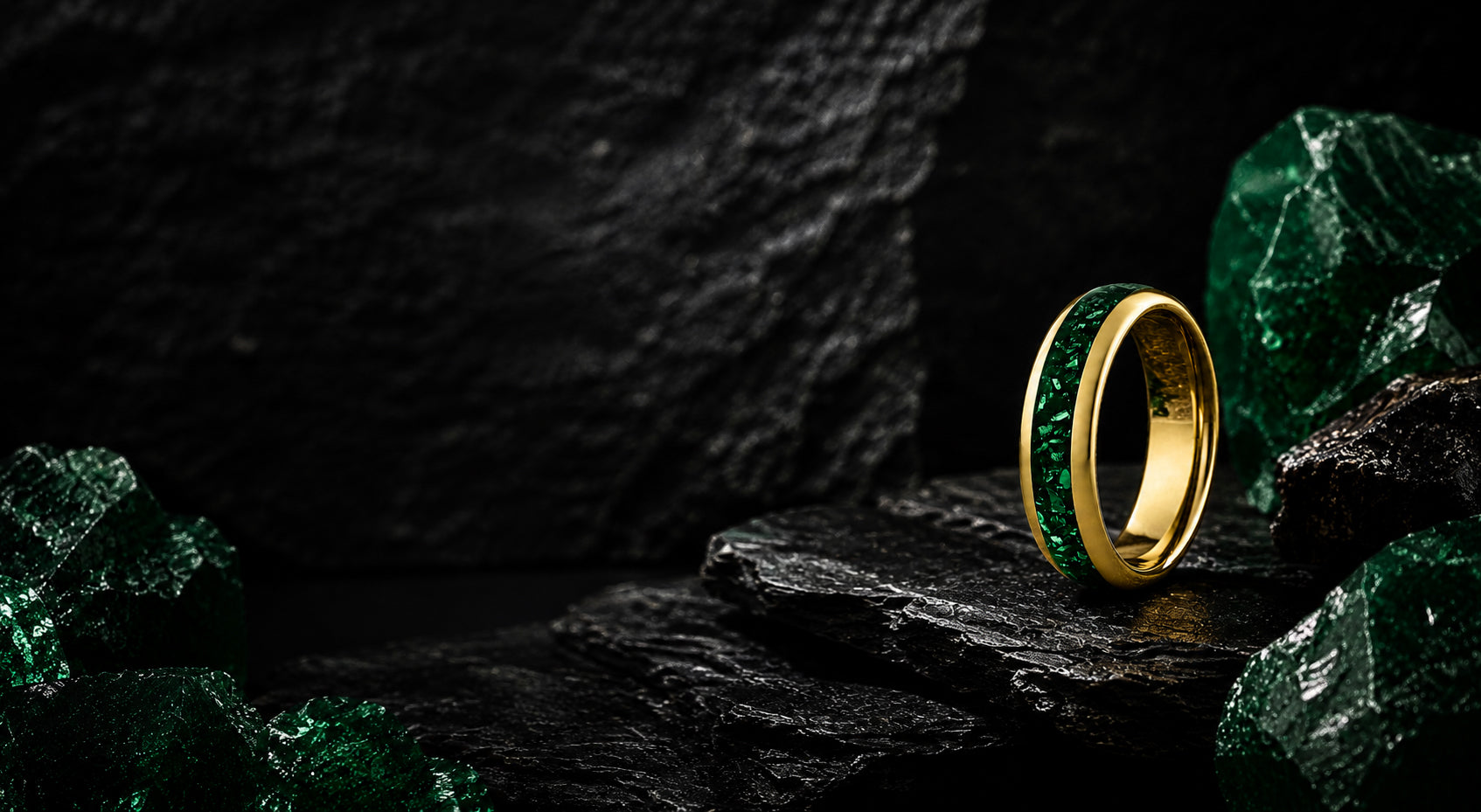 Wide desktop banner for emerald wedding rings collection featuring a gold tungsten ring with green emerald gemstone inlay positioned on a dark luxury slate background.