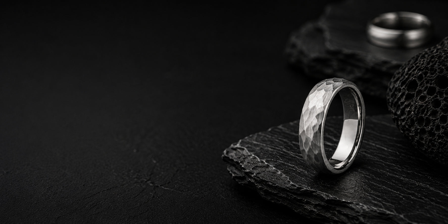 Hammered silver tungsten wedding band on dark volcanic rock with moody cinematic lighting and rugged charcoal slate textures