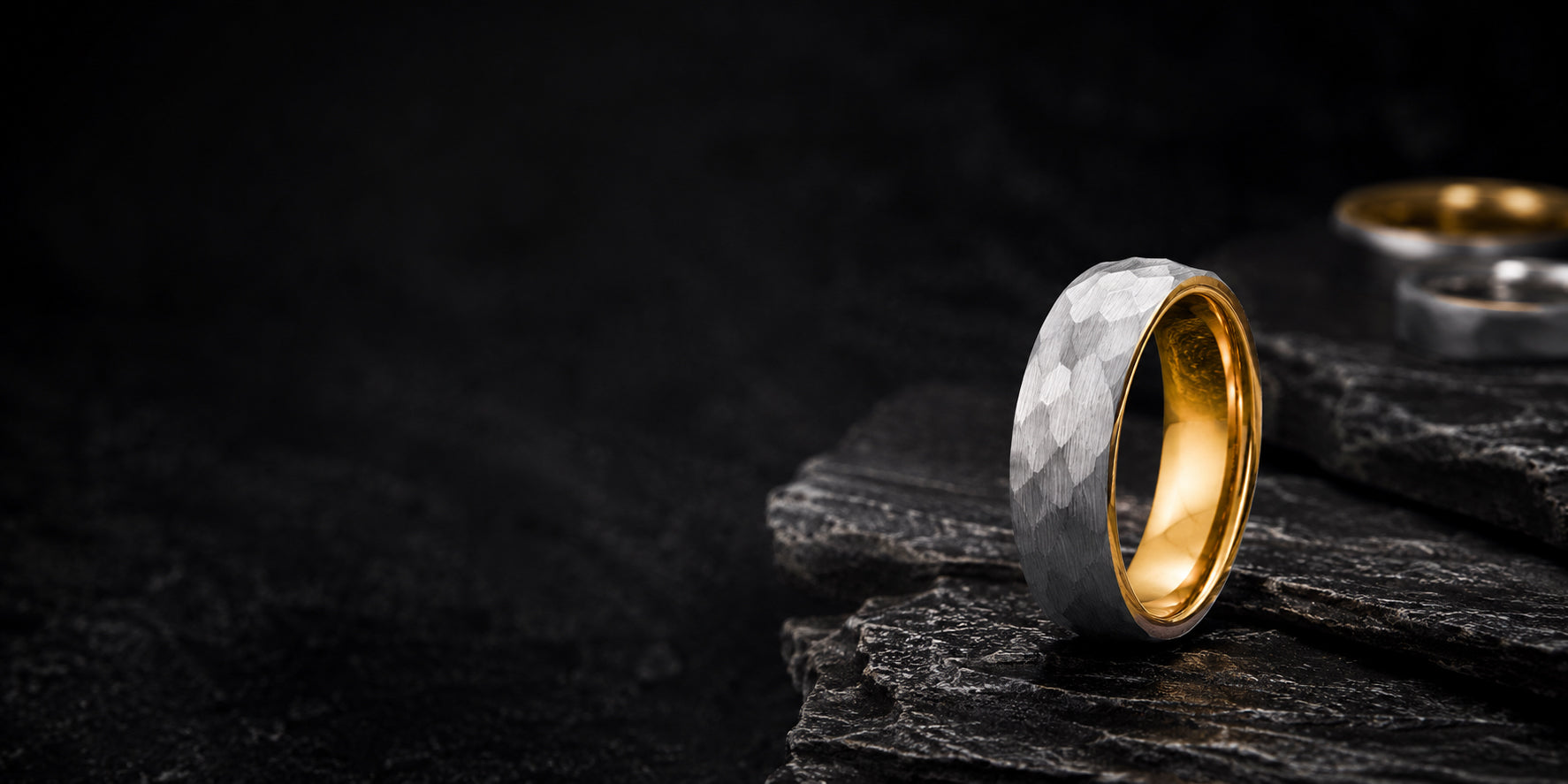 Hammered silver and gold men’s wedding ring on dark volcanic rock with moody cinematic lighting and rugged slate textures.
