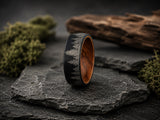 Black men’s wedding ring with engraved forest design and warm wood interior displayed on dark volcanic rock in a premium cinematic ecommerce hero scene.
