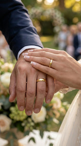 Two hands holding each other with wedding rings, surrounded by greenery and people in the background.