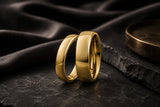 Matching polished gold wedding bands on a dark luxury textured background with warm cinematic lighting