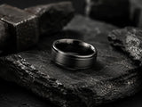 Dark cinematic product image of a durable black tungsten wedding ring resting on rugged slate stone textures with masculine industrial styling.
