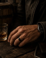Moody cinematic lifestyle image of a man wearing a hammered black wedding ring beside whiskey and dark wood textures with rugged masculine styling.