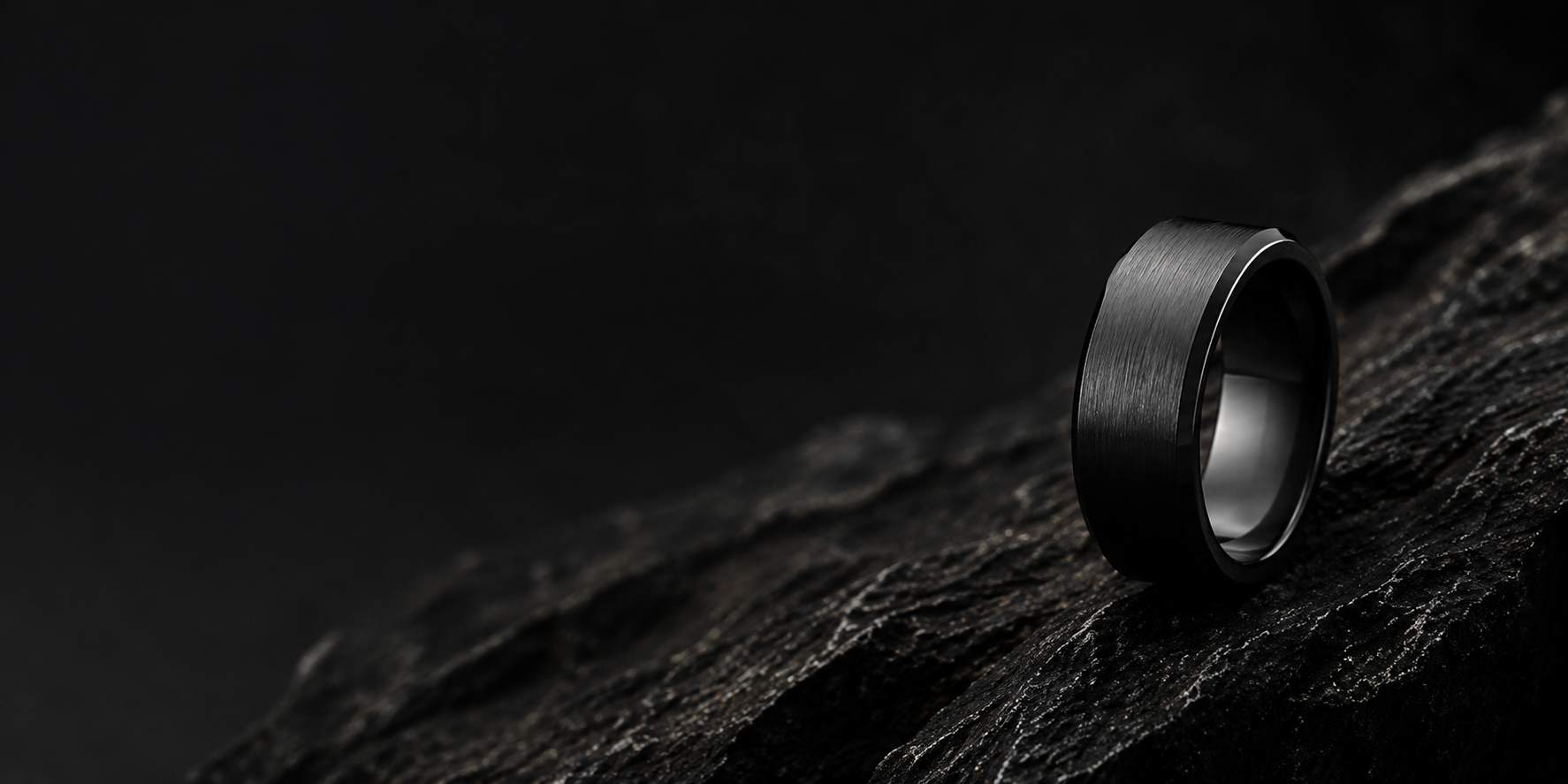 Men’s wedding bands collection hero image featuring a matte black tungsten ring on dark volcanic stone.