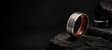 Luxury men’s wedding ring hero banner featuring brushed silver and whiskey barrel wood ring against dark charcoal stone background with cinematic lighting