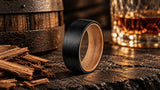 Black tungsten whiskey barrel wedding ring with warm natural wood interior in a rustic setting